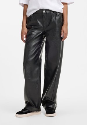 Persona che indossa pantaloni larghi in ecopelle nera e sneaker nere con suola bianca, in piedi davanti a uno sfondo semplice.