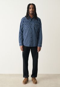 Chemise en flanelle à carreaux bleu marine avec deux poches poitrine, fermeture à boutons et manches longues, associée à un pantalon noir et des chaussures marron.