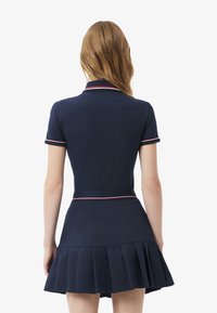 Donna con lunghi capelli castano chiaro che indossa un vestito blu navy a maniche corte con gonna plissettata e bordi a righe rosse e bianche, vista di spalle.