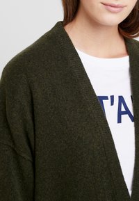 Olive green cardigan with open front, made of soft knit material. Features a wide collar and relaxed fit, layered over a white t-shirt.