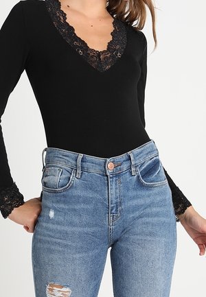 Femme portant un haut noir à manches longues avec un bord en dentelle au niveau du col et des poignets, associé à un jean bleu déchiré taille haute.