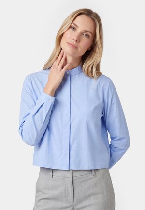 Frau mit blondem Haar, die ein hellblaues Hemd mit Knöpfen und graue Hose trägt, berührt mit der rechten Hand ihren Hals und posiert vor weißem Hintergrund.