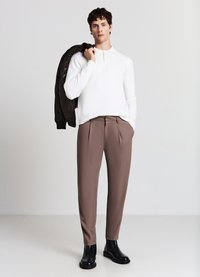 Weißes langärmliges Polohemd, braune gefaltete Hose und schwarze Stiefeletten. Modell hält eine dunkelbraune Jacke über einer Schulter.