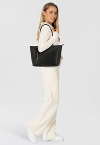 Sac en cuir noir avec une finition texturée, deux poignées, et un détail de frange décoratif, présenté aux côtés d'un mannequin portant une tenue claire.