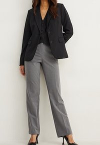 Femme portant un blazer noir et un gilet avec un pantalon gris ajusté et des escarpins noirs, se tenant devant un mur blanc.