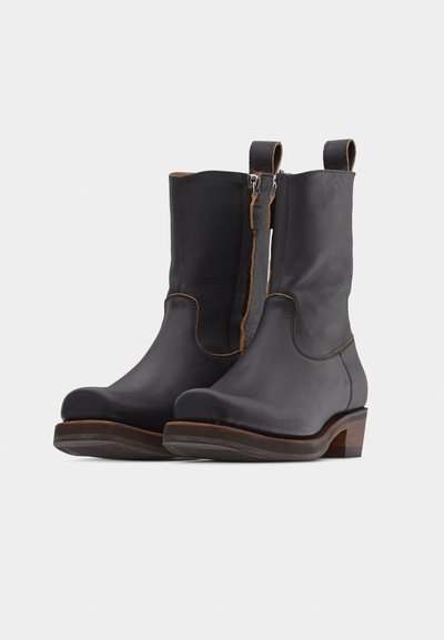 Μαύρες δερμάτινες μπότες μέχρι τη μέση της γάμπας με καφέ ραφές, πλαϊνά φερμουάρ, θηλιές για τράβηγμα και χαμηλά στρωμένα ξύλινα τακούνια σε λευκό φόντο.