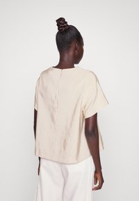 Beige lyhythihainen paita, jossa on pyöreä pääntie ja tekstuuri, takana napillinen suljenta. Yhdistetty vaaleisiin housuihin.