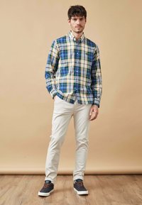 Camisa de cuadros en verde, azul y amarillo, con cuello abotonado. Combinada con pantalones beige claro y zapatillas oscuras con cordones naranjas.