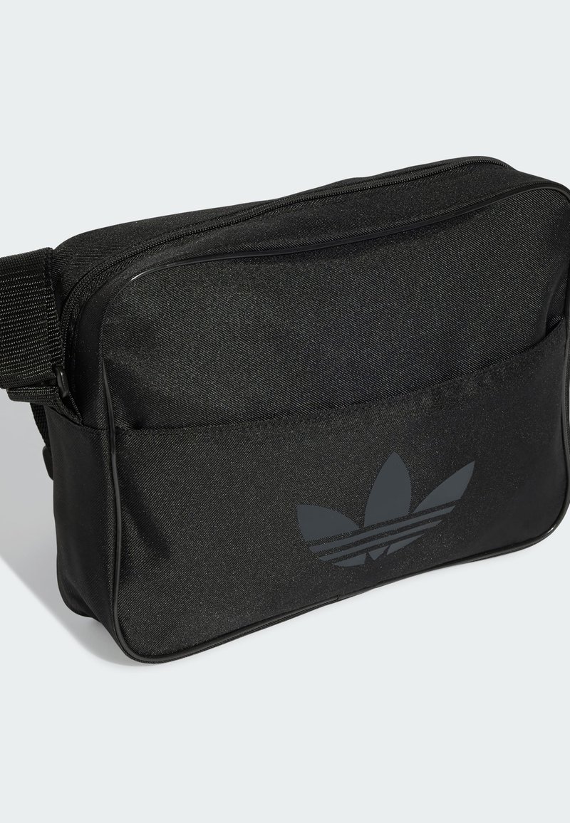 Sac en tissu noir avec fermeture éclair, arborant un logo Adidas gris discret et une surface texturée. Bandoulière réglable pour plus de confort.