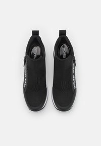 Pár černých slip-on tenisek Michael Kors s pletenými svršky, bočními zipy, bílými mezi-podrážkami a značkovými stélkami na bílém pozadí.