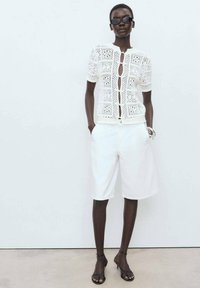 White crocheted knit top with short sleeves and button closure, paired with white shorts and black strappy sandals. Minimalist design.