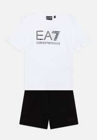 T-shirt blanc à manches courtes avec un logo EA7 noir, associé à un short noir portant une petite imprimé EA7 sur la jambe, fabriqué en coton lisse.
