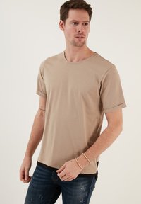 Beige Baumwoll-T-Shirt mit Rundhalsausschnitt und umgeschlagenen Ärmeln, mit einem schwarzen Saum und legerer Passform, getragen mit dunklen Jeans.