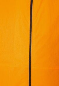 Orange jacka i tyg med en jämn textur har en central svart dragkedja och kontrasterande sömmar, vilket framhäver en ren, modern design.