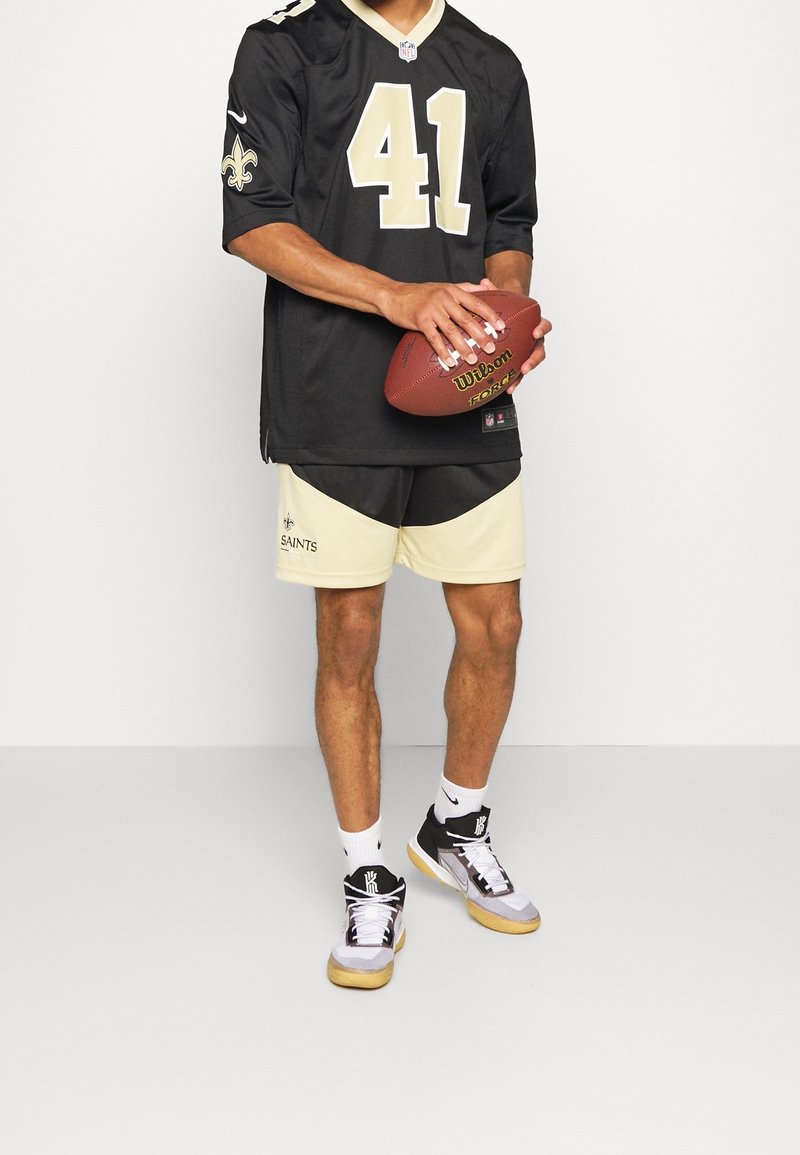 Personne portant un maillot et un short de football américain noirs et or des New Orleans Saints, tenant un ballon Wilson NFL marron sur un fond uni.