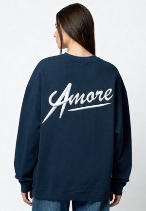 Donna con lunghi capelli scuri che indossa una felpa blu scuro con la scritta bianca grande "Amore" ricamata sulla schiena, girata di schiena.