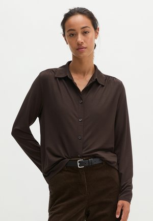 Vrouw met donker haar in een lage knot, draagt een donkerbruin overhemd met knopen, ingestopt in een bruine corduroy broek met een zwarte riem.