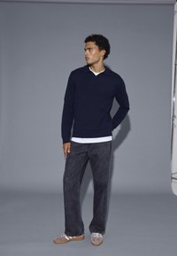 Marinefarbener Pullover mit V-Ausschnitt, über einem weißen Hemd getragen, kombiniert mit dunkelgrauen Hosen und grauen Turnschuhen.
