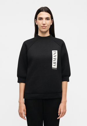 Femme aux cheveux lisses et foncés portant un sweat-shirt noir à manches mi-longues avec le logo vertical blanc "ARMANI EXCHANGE" sur le devant, debout devant un fond uni.