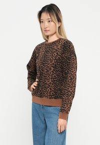 Sweater com padrão de leopardo castanho, mangas longas e bainha canelada. Feito de material suave, apresenta um decote redondo e corte descontraído. Usado com jeans azuis.