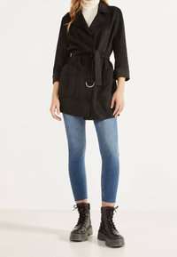 Trench coat in suede nero con vita sfilata, due tasche frontali e un accento in anello argentato. Indossato sopra un dolcevita crema e jeans blu.