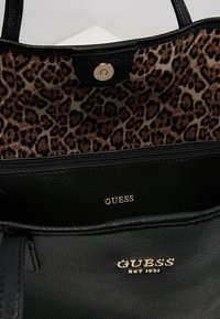 Sac à main Guess en cuir noir avec logo doré et doublure en tissu imprimé léopard visible à l'intérieur près de la fermeture magnétique.