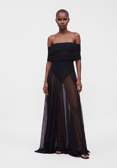 Femme aux cheveux courts portant un haut noir épaules dénudées et une longue jupe noire transparente par-dessus un short noir, debout devant un fond clair uni.