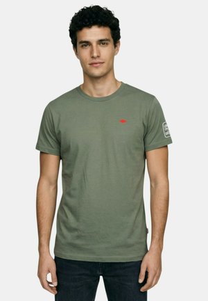 Jeune homme portant un t-shirt vert olive avec un petit logo rouge sur la poitrine et un motif blanc sur la manche gauche, debout devant un fond uni.