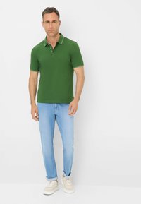 Polo verde con rifiniture bianche a contrasto, abbinato a jeans azzurri chiari e sneakers beige. Design semplice, maniche corte, vestibilità standard.