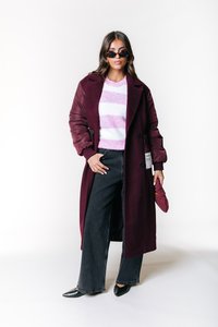 Un manteau long bordeaux avec des manches bouffantes, associé à un pull rayé blanc et rose. Un jean large noir et des chaussures noires à bout pointu complètent le look.