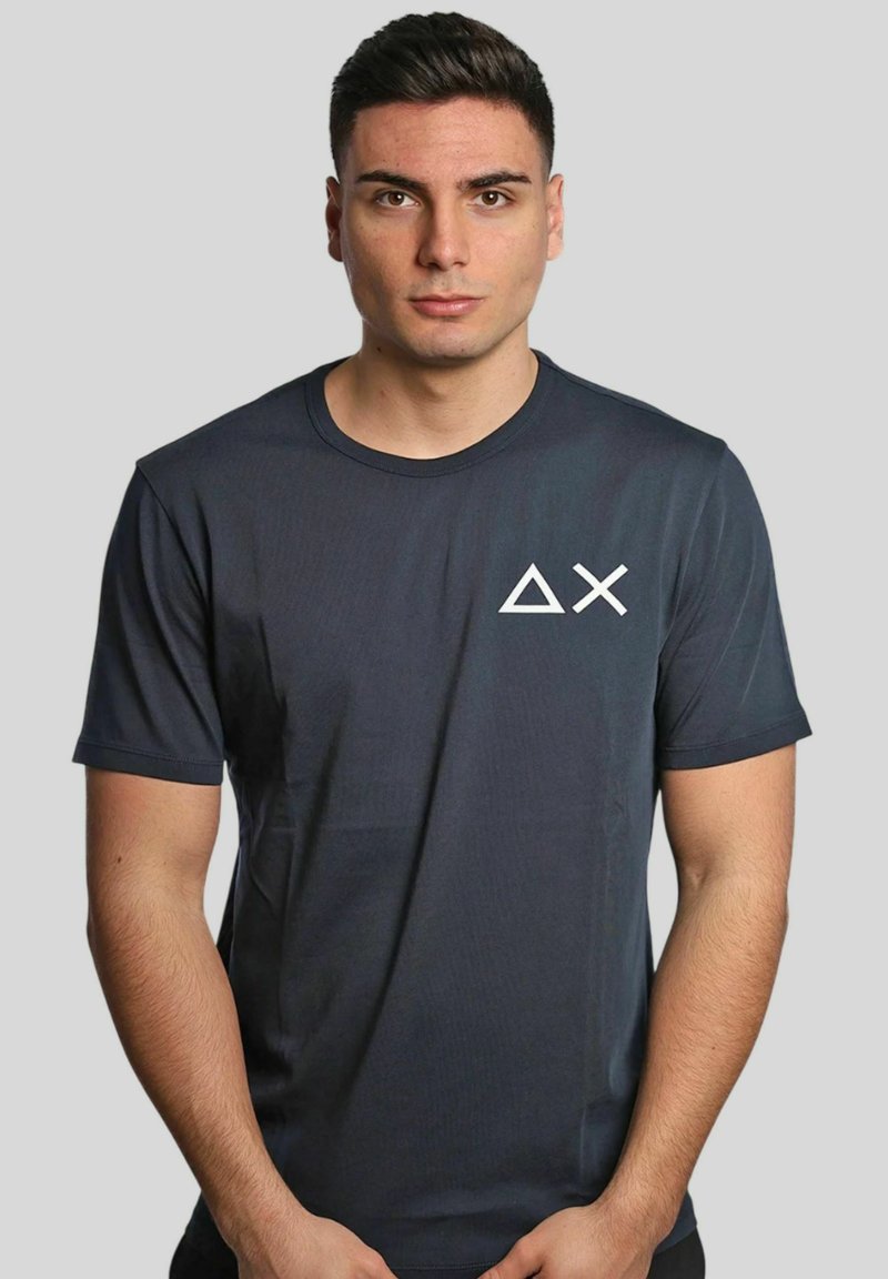 T-shirt corto grigio scuro realizzato in un tessuto liscio, con un logo geometrico bianco sul lato sinistro del petto. Design classico con collo a giro.