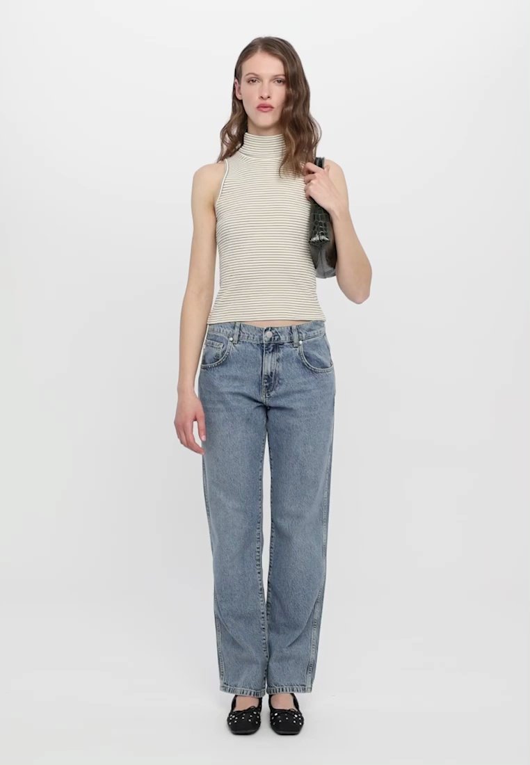 Haut sans manches rayé blanc et beige, jean taille haute bleu, et ballerines noires avec des clous ; modèle tenant une veste grise.