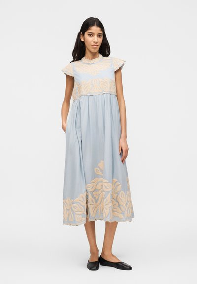 Γυναίκα με φόρεμα μέσης μήκους σε ανοιχτό μπλε χρώμα με μπεζ ανθικό κέντημα, μικρά μανίκια και μαύρα επίπεδα παπούτσια, που στέκεται μπροστά σε λευκό φόντο.