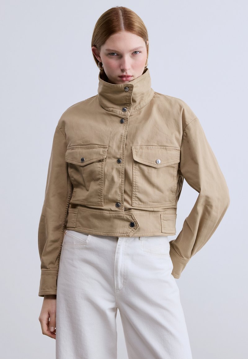 Κοντό μπεζ μπουφάν από μαλακό βαμβάκι, με ψηλό γιακά, δύο τσέπες στο στήθος με καπάκια και κλείσιμο με κουμπιά τύπου snap. Συνδυάζεται με λευκό παντελόνι.