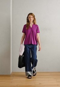 Violetti lyhythihainen paita napilla kaula-aukossa, yhdistettynä leveälahkeisiin denimpunttseihin, joissa on revitty helma, ja mustiin sandaaleihin. Kantaa vaaleanpunaista laukkua.