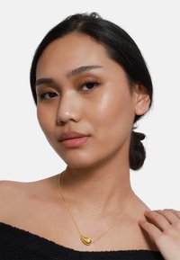 Goldene Halskette mit einem Tropfenanhänger an einer dünnen Kette. Das Model trägt ein schulterfreies Oberteil, das den Schmuck gut zur Geltung bringt.