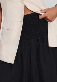 Gilet en lin beige avec un seul bouton et une poche, superposé sur une jupe smockée noire, avec un bas évasé.