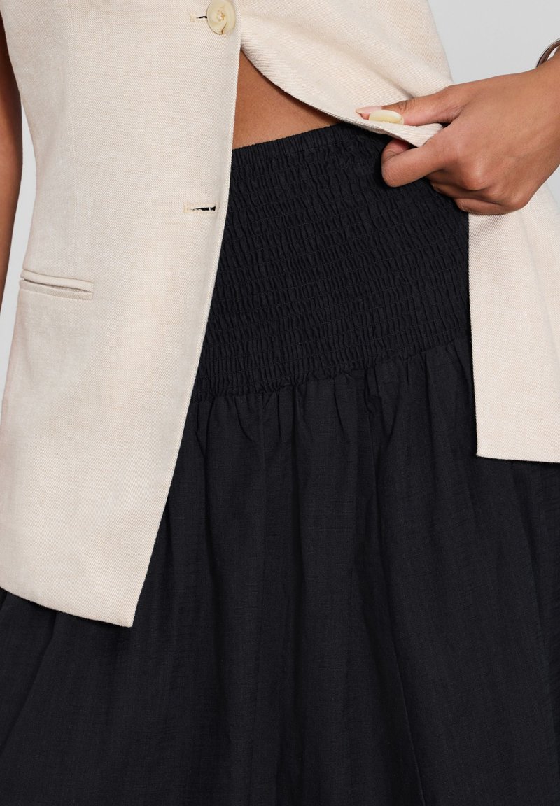 Gilet en lin beige avec un seul bouton et une poche, superposé sur une jupe smockée noire, avec un bas évasé.