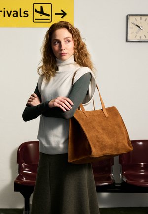 Junge Frau mit lockigem Haar steht mit verschränkten Armen und hält eine große braune Wildlederhandtasche im Wartebereich des Flughafens unter dem Ankunftsschild.