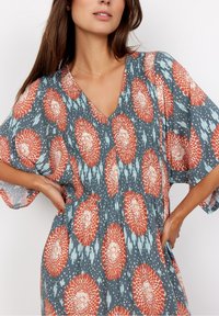 Leichtes Kleid mit floralem Muster in Türkis, mit orangefarbenen und weißen Blüten. V-Ausschnitt, kurze Ärmel und geraffte Taillenpartie.