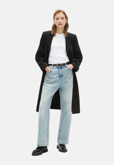 Un manteau noir long sur un t-shirt blanc, un jean droit bleu clair et des chaussures noires. La tenue présente une ceinture discrète et une coupe décontractée.