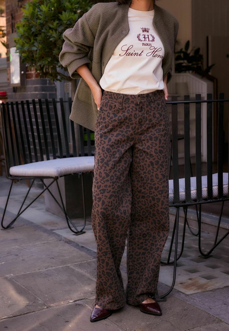 Personne portant un t-shirt graphique blanc, un cardigan vert olive et un pantalon large à motif léopard, debout sur un pavé en pierre près de chaises d'extérieur.
