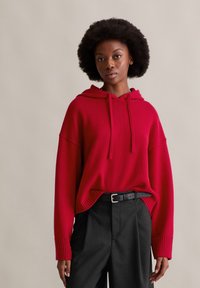 Felpa con cappuccio rossa in maglia con vestibilità comoda, dettagli a costine e cappuccio con coulisse, abbinata a pantaloni neri a righe sottili e cintura nera.