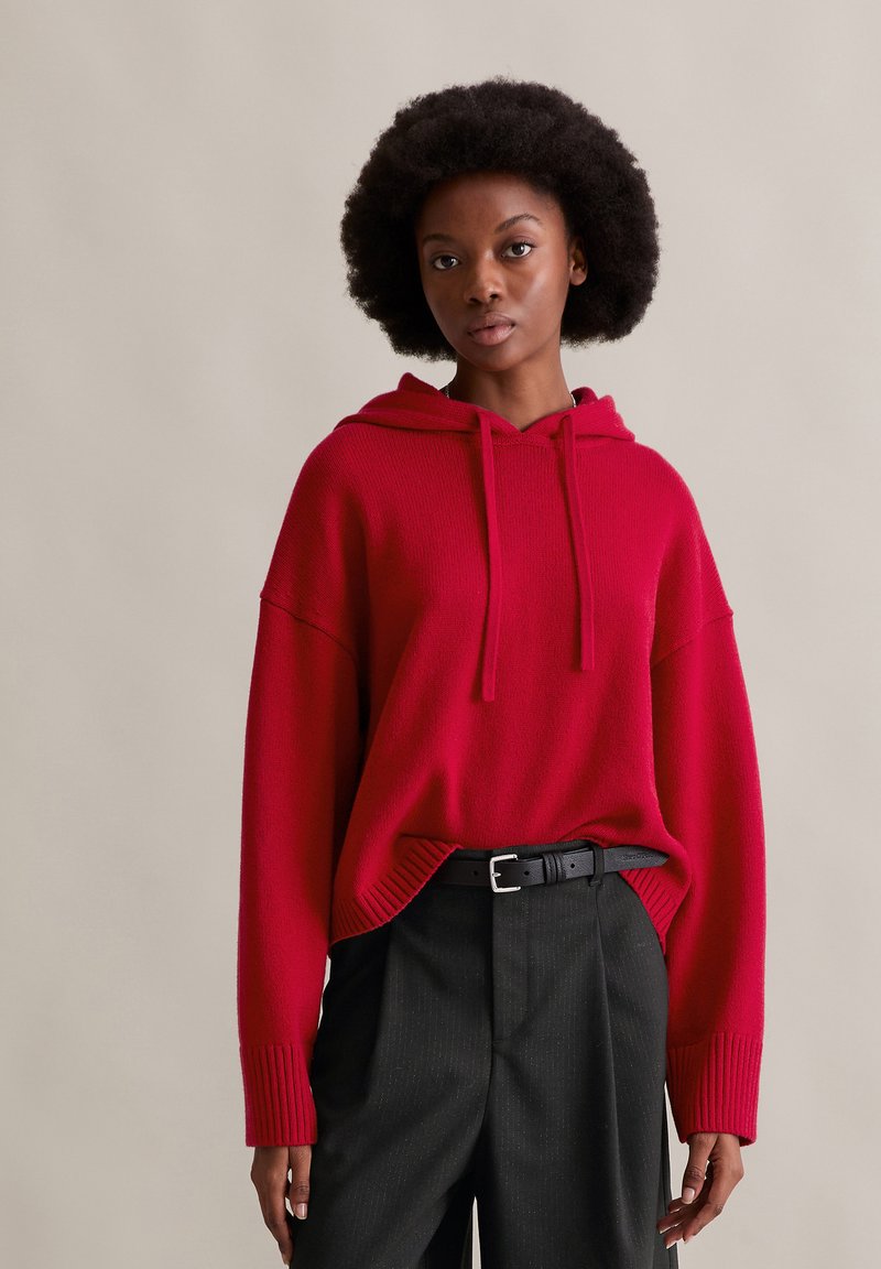 Felpa con cappuccio rossa in maglia con vestibilità comoda, dettagli a costine e cappuccio con coulisse, abbinata a pantaloni neri a righe sottili e cintura nera.