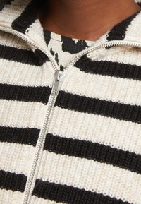 Gros plan sur un pull en maille rayé noir et blanc partiellement zippé sur un haut à motifs noir et blanc, porté sur une peau brune.