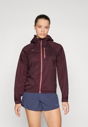 Ung kvinne iført en burgunderfarget hettejakke og marineblå treningsshorts, stående mot en ensfarget lys bakgrunn.
