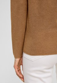 Pull en tricot marron avec un motif texturé, doté d'un ourlet côtelé et de manches longues, associé à un jean blanc.