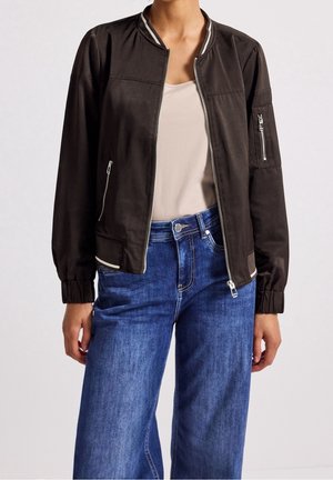 Mujer con chaqueta bomber negra con cremallera sobre un top beige combinada con vaqueros azules de tiro alto y pierna ancha, de pie frente a un fondo liso.