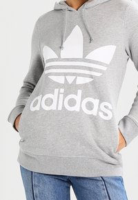 Person som bär en grå Adidas-hoodie med en stor vit logo och text, händerna i fickorna, tillsammans med blå jeans.