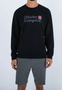 Muž v černé mikině s logem "The Hurley Company" a malým barevným emblémem, oblečený v šedých šortkách, stojí na bílém pozadí.